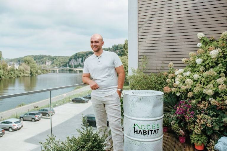 Man standing on a balcony beside a river, smiling, with potted plants and a metal trash bin that reads 'Accès Habitat'.