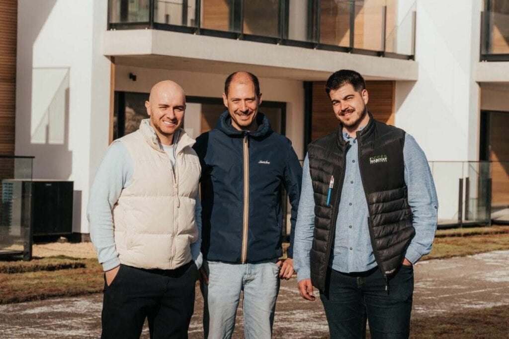 Three men stand outside a modern white building, smiling at the camera with hands in pockets.